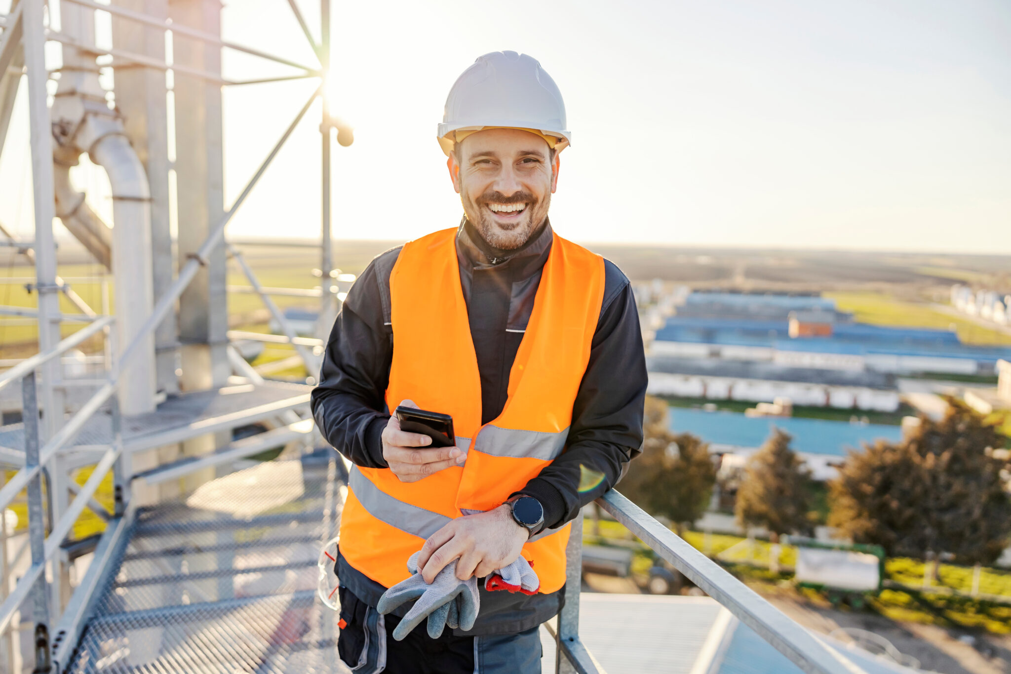 A happy industry worker holding phone on height on metal construction and smiling.