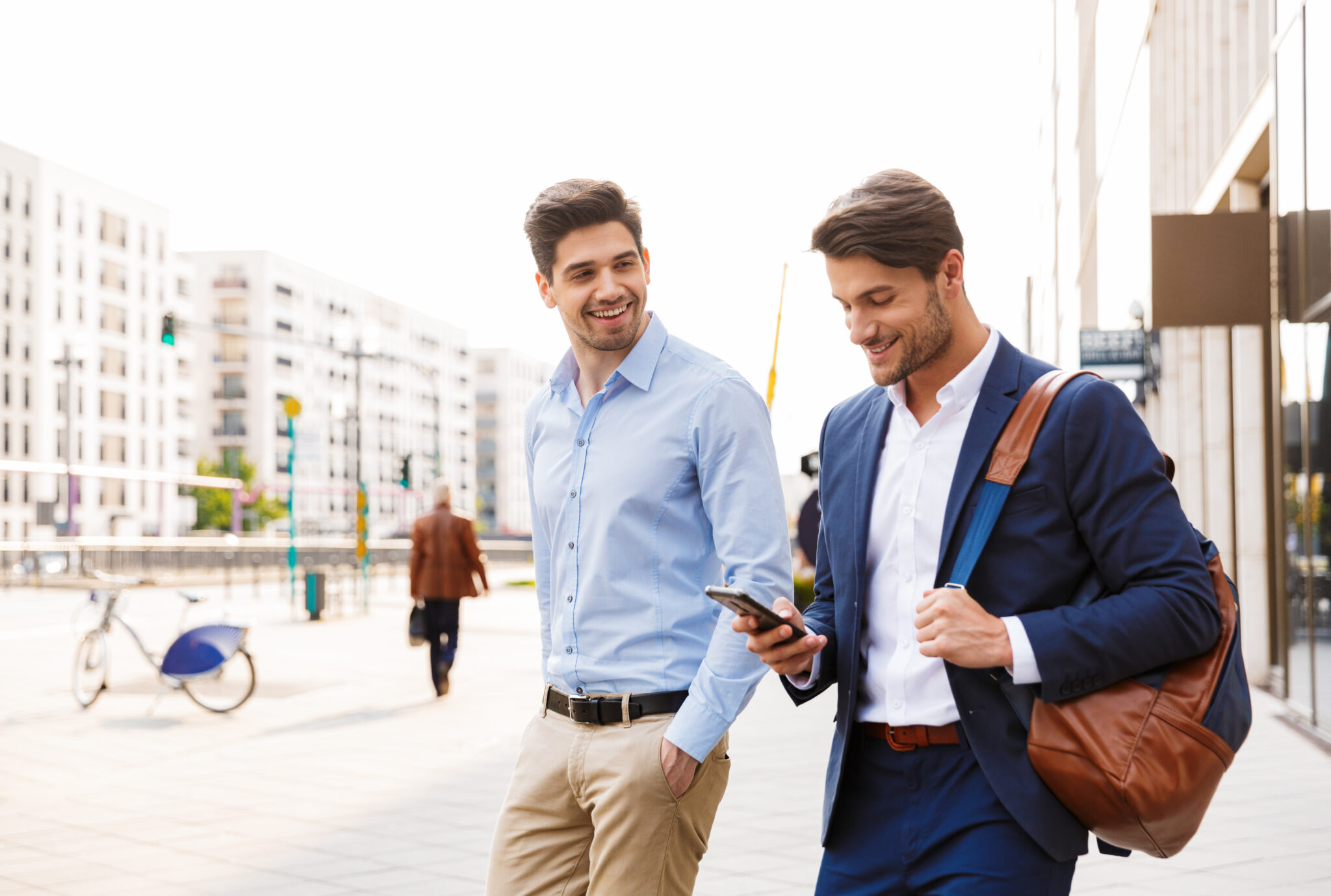 Image of a cheerful happy young two friends colleagues business men walking near business center talking with each other using mobile phone.