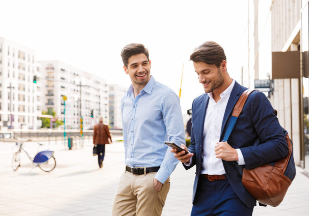 Image of a cheerful happy young two friends colleagues business men walking near business center talking with each other using mobile phone.