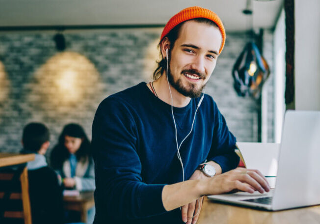 Portrait of happy smiling hipster guy enjoying time for favourite music playlist during e learning in cafeteria, positive male teenager in headphones rest indoors sitting at table with laptop device