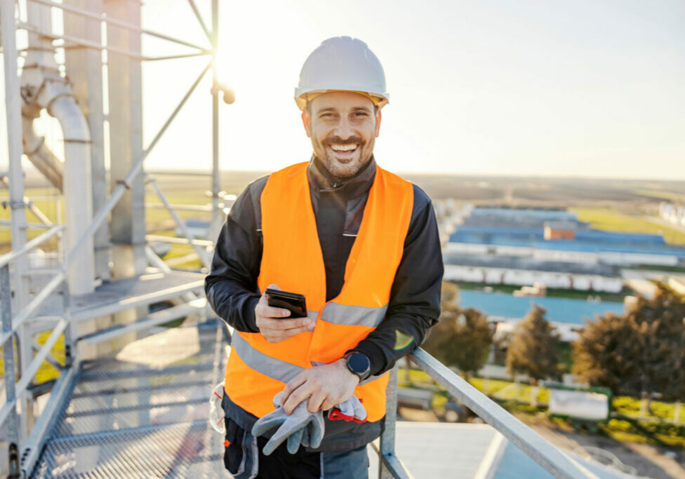 A happy industry worker holding phone on height on metal construction and smiling.